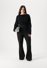 Pulover lung negru, cu mânecă lungă, și croială lejeră, asortat cu pantaloni negri la brâu înalt. Modelul ține o poșetă metalică argintie. Fundal simplu.