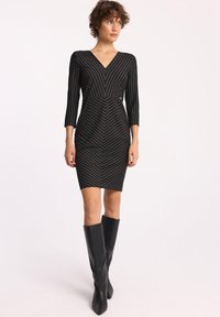 Czarna sukienka w pionowe i w kształcie chevron białe paski, dekolt w serek, rękawy do trzy czwarte, dopasowany krój, zestawiona z wysokimi czarnymi botkami.