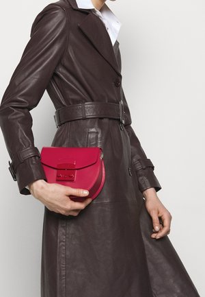 Vrouw draagt een donkerbruine leren trenchcoat en houdt een kleine rode leren handtas met voorste sluiting vast tegen een witte achtergrond.