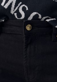 Jeans de mezclilla negra con cierre de botón dorado. Incluye bucles para cinturón y costuras laterales, con una tela texturizada y sin patrones visibles.