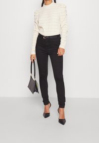 White, textured long-sleeve top with puffed shoulders, paired with black skinny jeans and black pointed-toe heels. Small black handbag with handle.