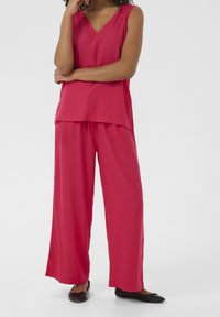 Haut rouge sans manches à col en V, associé à un pantalon ample à jambes larges. Tissu lisse avec un léger éclat. Chaussures plates noires portées.