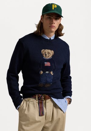 Maglione blu navy con un design a maglia di un orso, caratterizzato da una bandiera americana, abbinato a pantaloni khaki e una cintura a strisce multicolore. Cappellino verde.