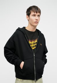 Must mustaga, millel on ees tasku, valmistatud pehmest kangast. Omab reguleeritavat kapuutsi ja tagasihoidlikku logodetaili.