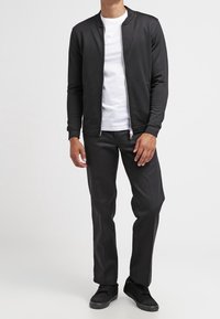 Veste bomber noire zippée portée sur un t-shirt blanc, associée à un pantalon foncé et des baskets noires. Texture lisse et design minimaliste.