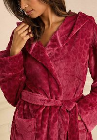 Fleece bathrobe in deep magenta with a textured pattern, featuring a hood, wide sleeves, a tie belt, and patch pockets. Soft fabric.