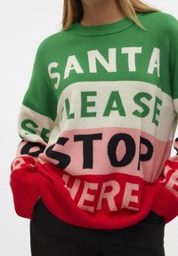 Pull avec des blocs de couleurs vert, blanc, rose et rouge portant un texte en gras disant "Santa Please Stop Here", porté par une personne les mains dans les poches.