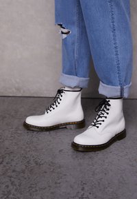Botas de combate de cuero blanco con cordones negros, costuras amarillas y una suela gruesa, combinadas con unos jeans azul claro, arremangados y con un desgarro.