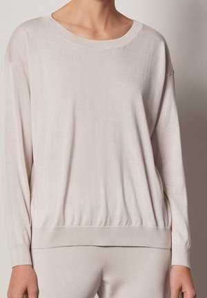 Persoon draagt een losse, beige trui met lange mouwen met geribbelde boorden en zoom, tegen een effen grijze achtergrond.