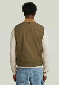 Giacca senza maniche verde oliva con colletto a coste, bottoni laterali e dettagli di cucitura. Indossata sopra un maglione crema e jeans blu.