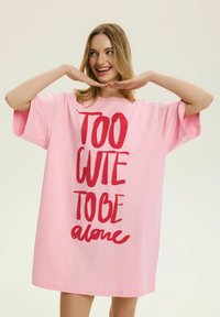 Uśmiechnięta kobieta w luźnej różowej sukience z czerwonym napisem „ZBYT SŁODKA, BY BYĆ sama”, trzymająca dłonie pod brodą na jednolitym tle.