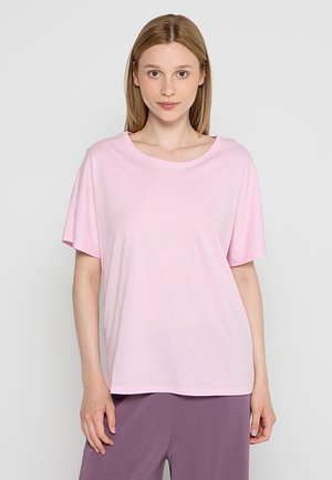 Młoda kobieta ubrana w luźny jasnoróżowy T-shirt i fioletowe spodnie, stojąca i zwrócona przodem na jednolitym tle.