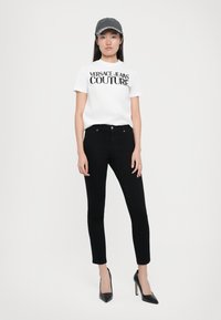 Λευκό μπλουζάκι με την επιγραφή "Versace Jeans Couture" σε μαύρο χρώμα, συνδυασμένο με εφαρμοστά μαύρα τζιν και γυαλιστερές μαύρες γόβες. Ένα γκρίζο καπέλο ολοκληρώνει το ντύσιμο.