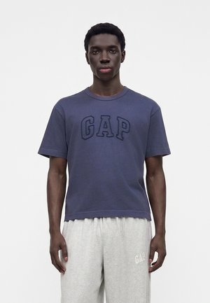 Ung mand iført en marineblå GAP T-shirt og lysegrå GAP joggingbukser står foran en ensfarvet hvid baggrund.