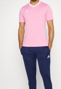 Maglietta atletica rosa con scollo a V e bordi bianchi, realizzata in tessuto traspirante, abbinata a pantaloni della tuta blu navy; entrambi presentano il logo Adidas.
