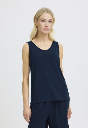 Femme aux cheveux blonds portant des boucles d'oreilles en or et un haut sans manches bleu marine avec un pantalon assorti, debout devant un fond blanc uni.