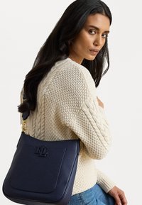 Ναυτικό δερμάτινο τσάντα ώμου με αυστηρή σιλουέτα, ανάγλυφο λογότυπο και χρυσά εξαρτήματα, φορεμένη πάνω από ένα πλεκτό κρεμ πουλόβερ.