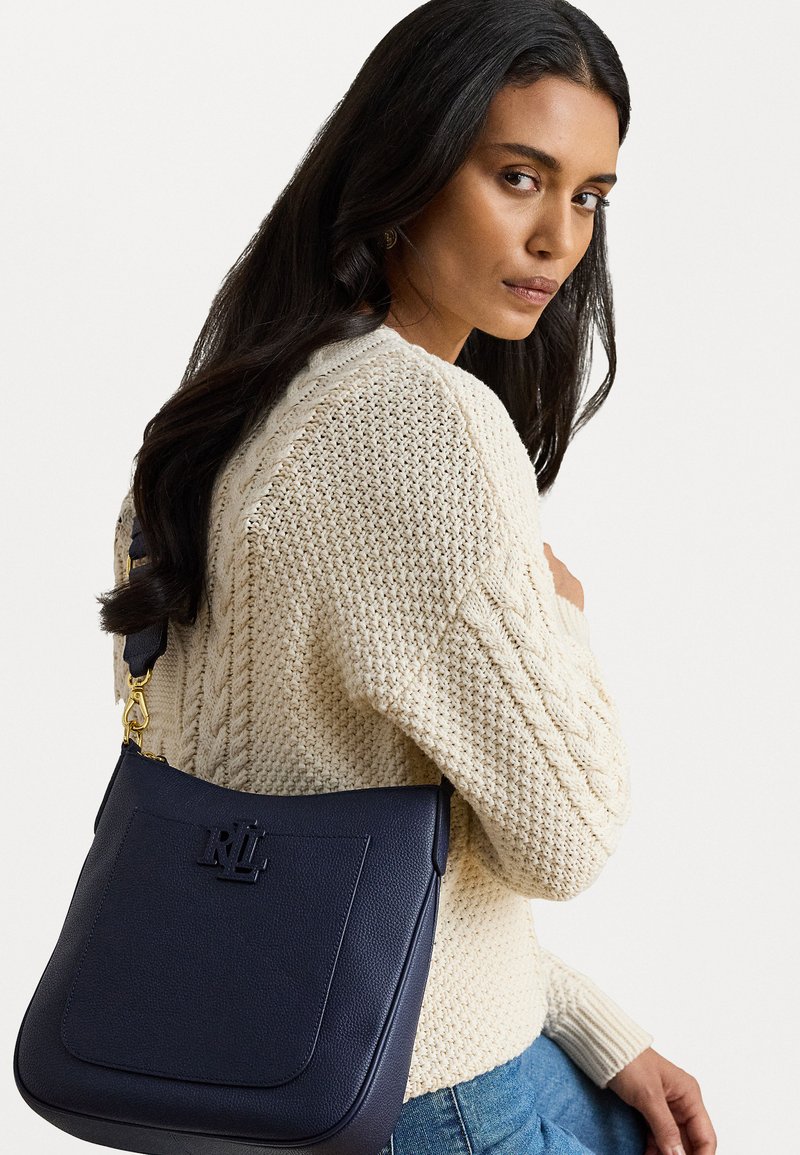 Ναυτικό δερμάτινο τσάντα ώμου με αυστηρή σιλουέτα, ανάγλυφο λογότυπο και χρυσά εξαρτήματα, φορεμένη πάνω από ένα πλεκτό κρεμ πουλόβερ.