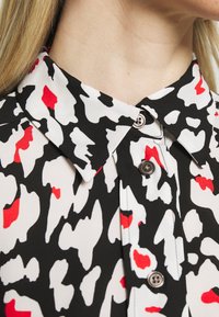 Gros plan du col d'une chemise à boutons avec un motif abstrait noir, blanc et rouge porté par une personne aux cheveux blonds.