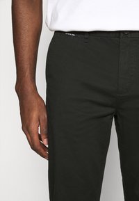 Pantalons noirs en coton à coupe droite, affichant une étiquette à la taille, des poches latérales et de subtils détails de couture. Tissu texturé.