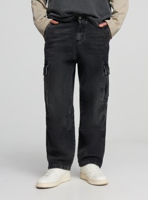 Pantaloni cargo in denim nero ampio con multiple tasche laterali, dettagli consumati e taglio a gamba dritta, abbinati a sneakers bianche.