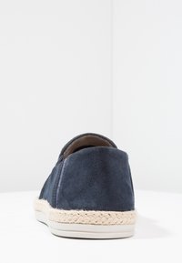 Chaussure slip-on en suède marine avec une semelle intermédiaire en jute tressé et une semelle en caoutchouc lisse. Design minimaliste sans quincaillerie visible.