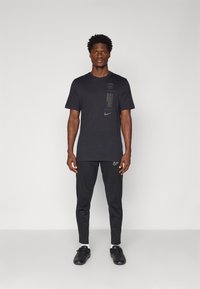 Černé tričko Nike s logem a textem, černé kalhoty Nike a černé boty. Materiál je hladký a má střih přiléhající k tělu.