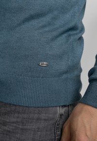 Turquoise trui met ronde hals van ribgebreide stof. Bevat een klein ovaal metalen logo-tag aan de onderkant. Draagt over een donkere broek.