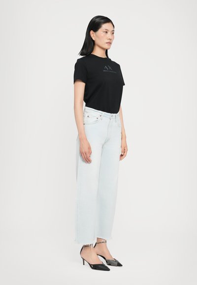 Μαύρο βαμβακερό μπλουζάκι με λογότυπο, ανοιχτό μπλε τζιν παντελόνι με ψηλή μέση και φαρδύ πόδι, και μαύρα πέδιλα slingback με μυτερά δάχτυλα και λουράκια στον αστράγαλο.