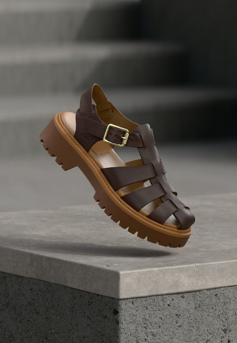 Brown leather sandal with chunky sole and gold buckle, balanced on edge of gray stone surface with blurred steps in background.