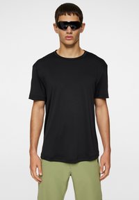 Svart kortärmad t-shirt med rund halsringning. Slät textur och avslappnad passform. Buren med gröna shorts, modellen har också svarta solglasögon.