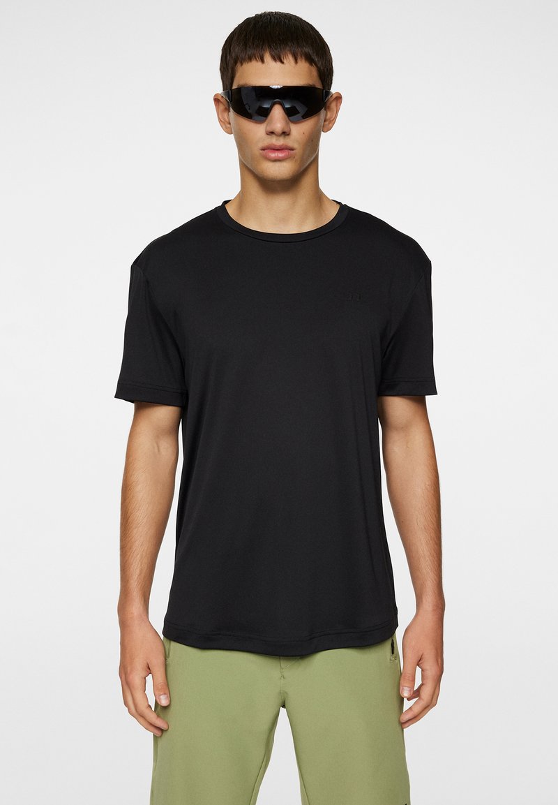 Svart kortärmad t-shirt med rund halsringning. Slät textur och avslappnad passform. Buren med gröna shorts, modellen har också svarta solglasögon.