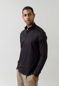 Cămașă neagră cu mâneci lungi, din bumbac, cu guler structurat, textură netedă și butoniere în față. Asortată cu pantaloni khaki de culoare deschisă.