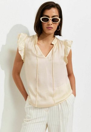 Donna con top crema senza maniche con volant, pantaloni bianchi a righe sottili, occhiali da sole ovali bianchi e orecchini a cerchio dorati, in piedi con le mani in tasca.