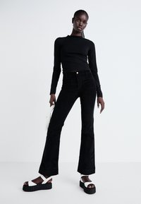 Svart ripset langermet topp kombinert med høyt liv svarte kordfløyels flared jeans. Modellen har på seg hvite platå-sandaler og holder en liten hvit veske.