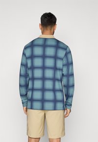 Μπλούζα μακρύ μανίκι με μπλε γραδιέ ν και σχέδιο πλέγματος, με μπεζ γιακά. Φορεμένη με μπεζ σορτς, που δείχνει λεία υφή υφάσματος.