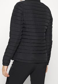 Svart puffjacka med horisontella quiltade sektioner, hög krage och ribbad mudd. Tyget verkar slätt och lätt.