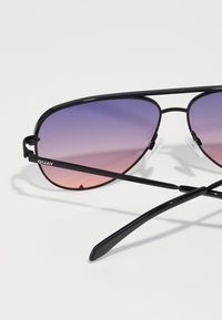 Schwarze Flieger-Sonnenbrille mit verlaufenden lila-zu-pinkfarbigen Gläsern, ausgestattet mit einem dünnen Metallrahmen und klaren Nasenpolstern. Markenname "QUAY" an der Seite.