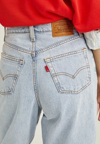 Vaaleansiniset farkut, joissa on takataskut, oranssi tikkaus ja ruskea nahkalamina, jossa on bränditiedot. Huomaa pieni punainen lippu sauman kohdalla.