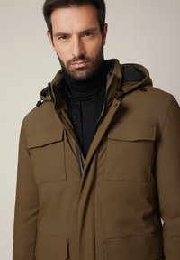 Brązowa kurtka z kapturem, zapinana na zamek z przodu, z dużymi kieszeniami na piersi i gładką fakturą. Nosi się na czarnym golfie.