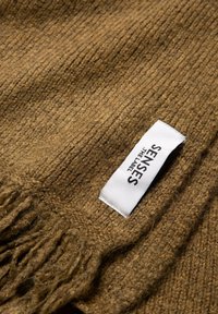 Close-up of brown ribbed knit fabric with fringes and a white label reading "SENSES THE LABEL" sewn on the edge.