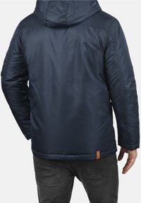 Uomo che indossa una giacca a zip con cappuccio di colore blu navy e jeans scuri, visto di spalle su uno sfondo grigio chiaro.