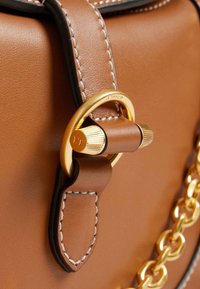 Dettaglio di una borsa in pelle marrone che mostra una chiusura in metallo dorato, una superficie liscia, bordi neri, cuciture bianche a contrasto e un'accento in catena.