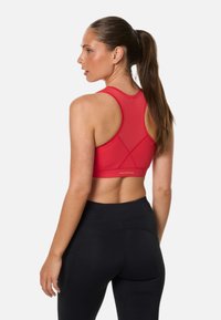 Kvinde med langt hår i en hestehale iført en rød racerback sports-bh og sorte højtaljede leggings, stående foran en hvid baggrund.