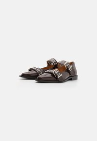 Zapatos de charol marrón con punta afilada, que presentan dos tiras anchas adornadas con acentos circulares y hebillas. Tacón bajo tipo bloque.