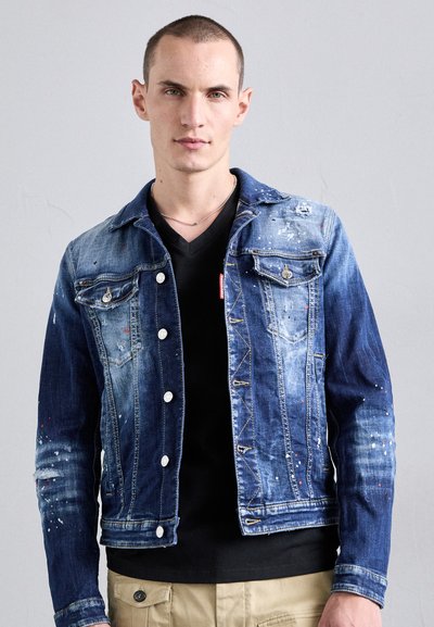Chaqueta de mezclilla azul deslavada con áreas desgastadas y salpicaduras de pintura blanca, con botones y bolsillos frontales. Camiseta negra de escote en V por debajo.