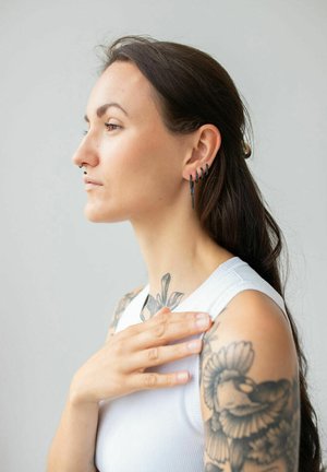 Black metal earrings with angular designs and a textured surface, worn with a sleeveless white top. Visible tattoos on the shoulder.