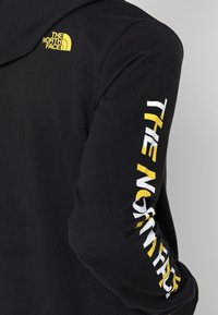 Schwarzer Kapuzenpullover mit einem gelben und weißen Logo auf Brust und Ärmel. Weicher Stoff, Standardpassform, mit klassischem Kapuzen-Design.