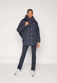 Námornícka puffer bunda so zlatými cvočkami a vysokým golierom; skombinovaná s tmavými nohavicami a sivými teniskami; model nesie čierny batoh.