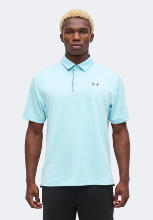 Mann mit kurzen lockigen blonden Haaren, der ein hellblaues Under Armour Kurzarm-Poloshirt und schwarze Hosen trägt, steht und schaut nach vorne.